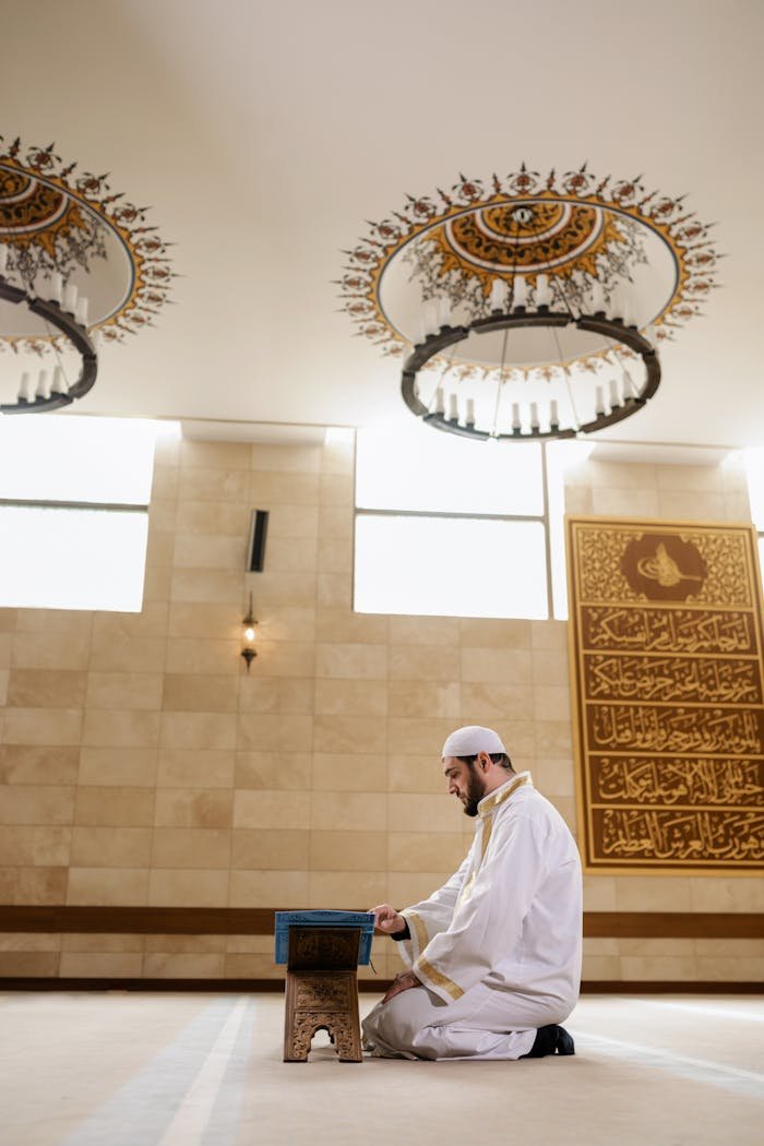 A Man in White Thobe Sitting on the Floor while Reading a Koran